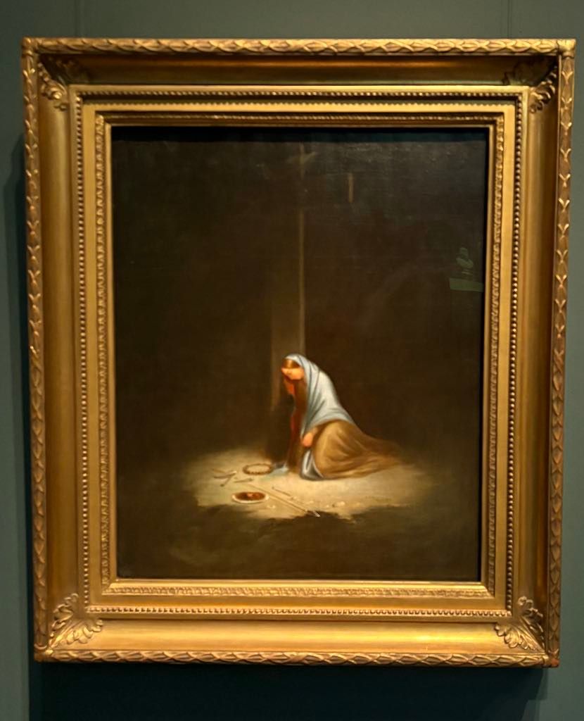 A framed painting depicts a kneeling woman in a white and brown robe with a headscarf, sitting on the floor in dim light beside two clay vessels.