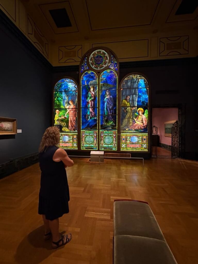A person stands in an art gallery looking at large, colorful stained glass panels depicting various figures and scenes, illuminated against a dark wall.