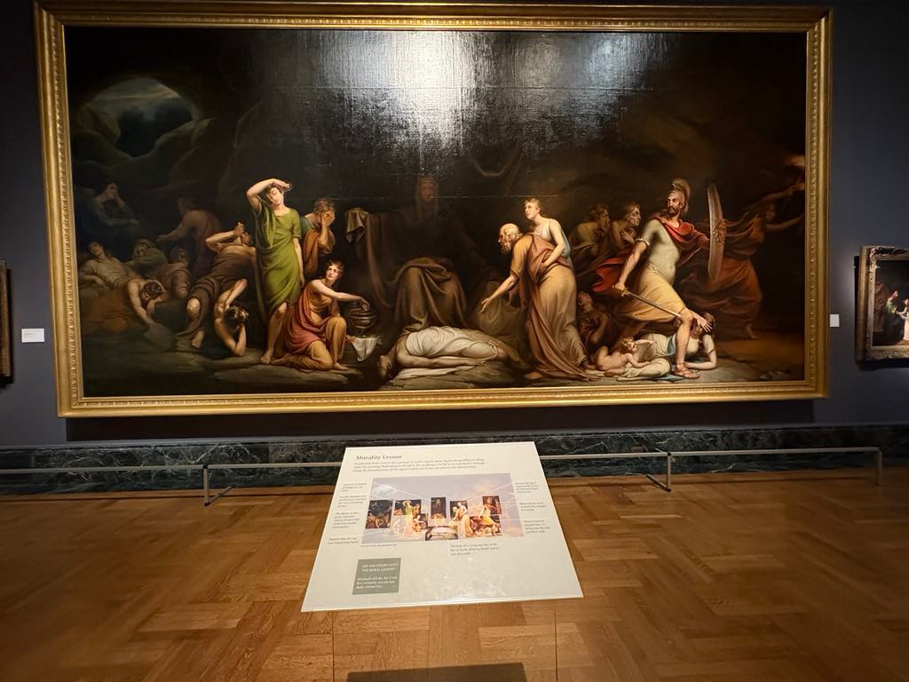 Large classical painting of a dramatic scene with several figures in a dark setting displayed in a museum, accompanied by an informational placard in front.
