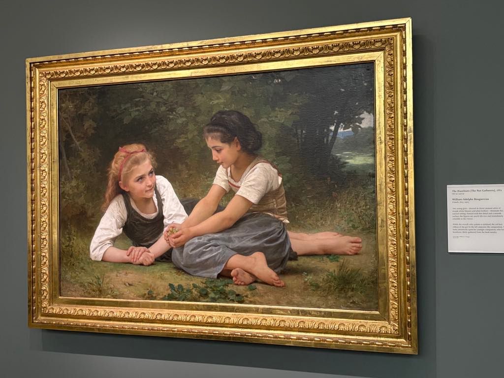 A framed painting of two young girls sitting on grass, one lying on her stomach and the other seated cross-legged, engaged in conversation under trees.