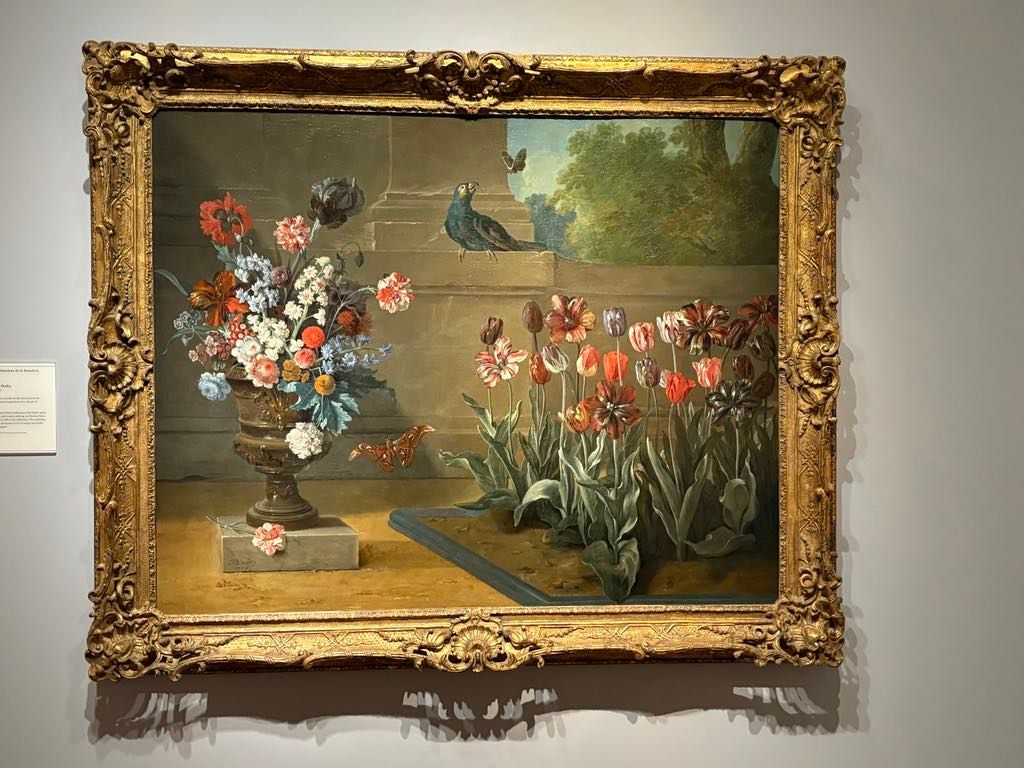 A framed painting depicting a bouquet of flowers in a vase and blooming tulips in a garden, with a bird and butterfly in the background.