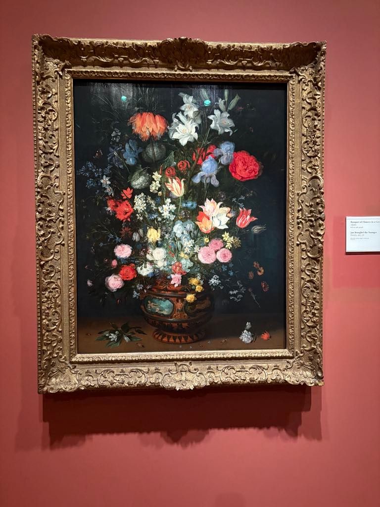 A framed still life painting of a bouquet of flowers in a decorative vase, displayed on a red wall with a small information plaque to the right.