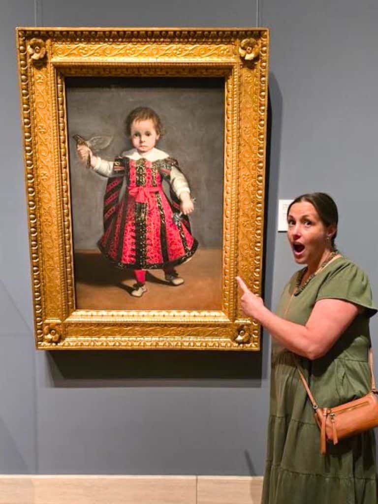 A woman in a green dress stands next to a framed painting of a child in ornate clothing, pointing at it with a surprised expression.