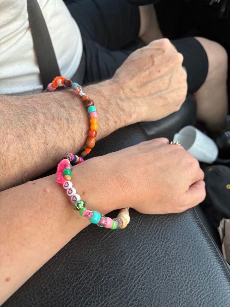 Two hands resting on a car armrest, each wearing a colorful beaded bracelet. The person in the front wears shorts, and a white travel mug is visible nearby.