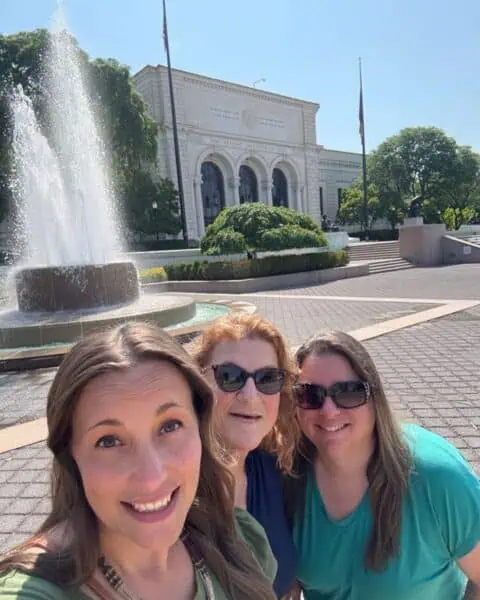 Three women smile for a selfie in front of a fountain and the Detroit Institute of Arts building on a sunny day.