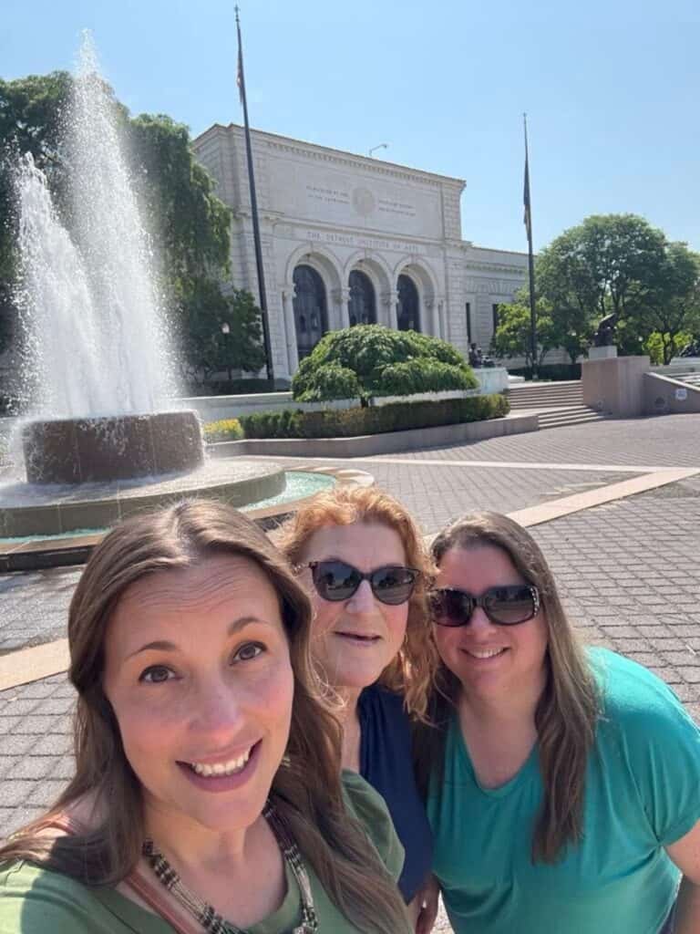 Three women smile for a selfie in front of a fountain and the Detroit Institute of Arts building on a sunny day.