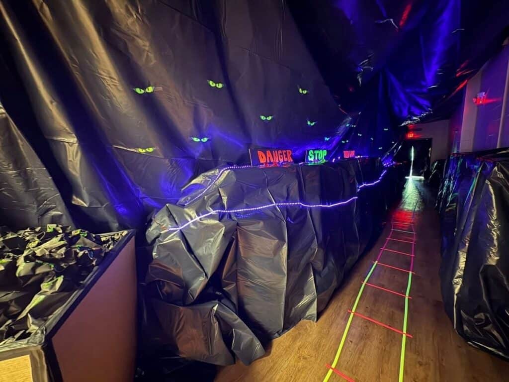 A hallway decorated with black plastic sheets, neon signs, glowing cat eyes, string lights, and neon tape on the floor, creating a spooky atmosphere.