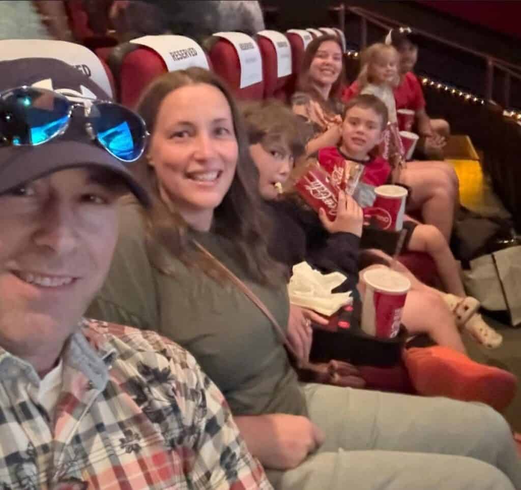 A group of people, including adults and children, sit in a movie theater with popcorn and drinks in their seats, smiling at the camera.