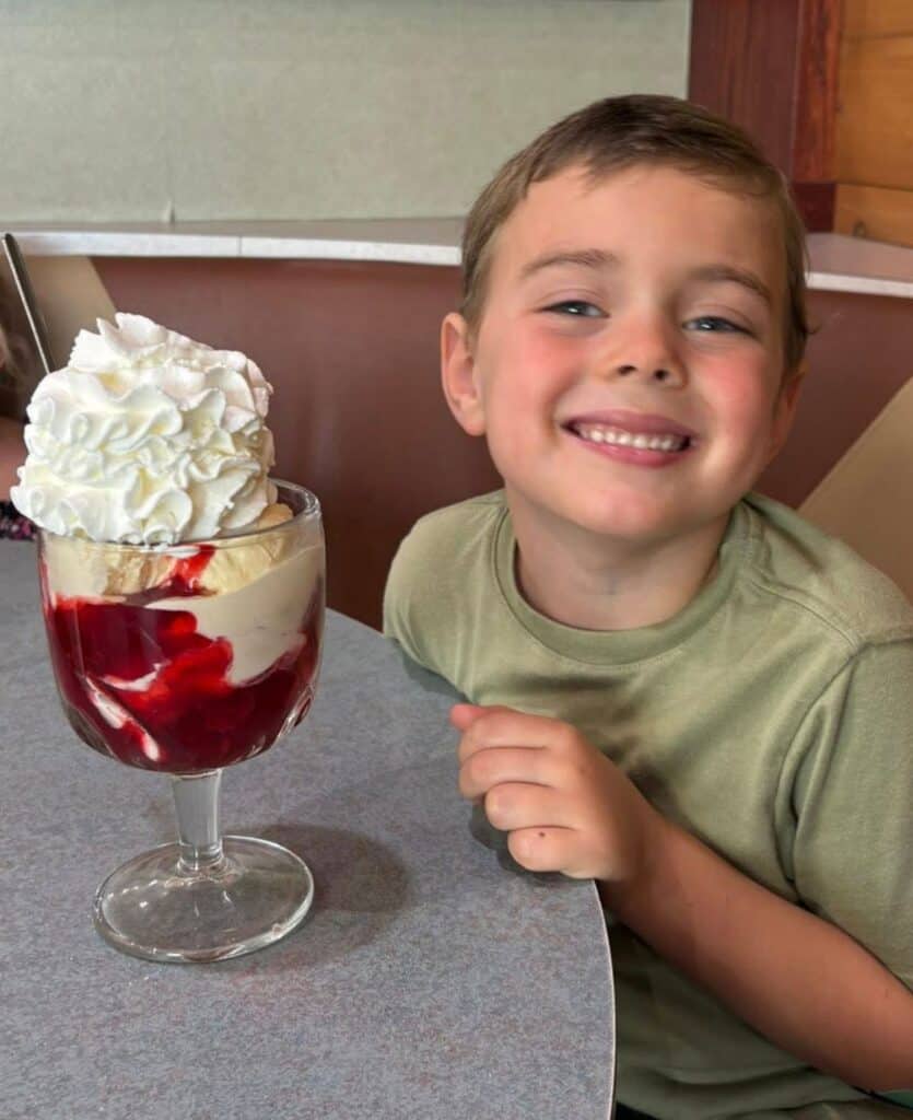A smiling child sits at a table next to a large glass dessert with whipped cream, vanilla ice cream, and strawberry sauce.