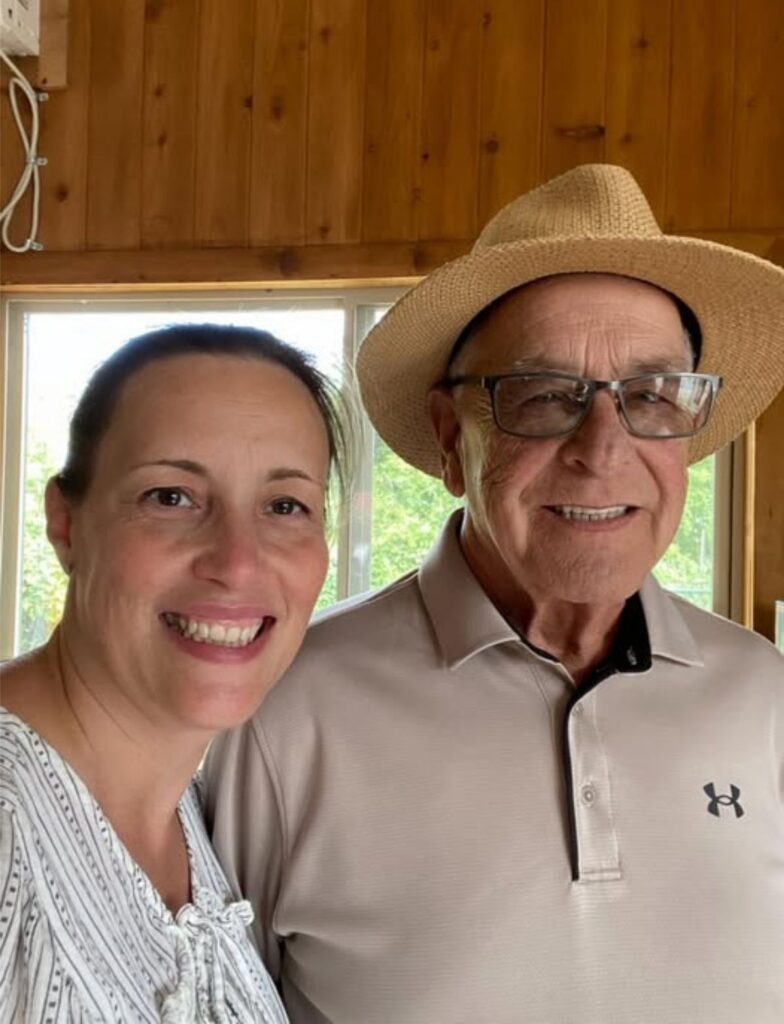 A smiling woman and an older man wearing glasses and a straw hat pose together indoors in front of a window and wooden wall.