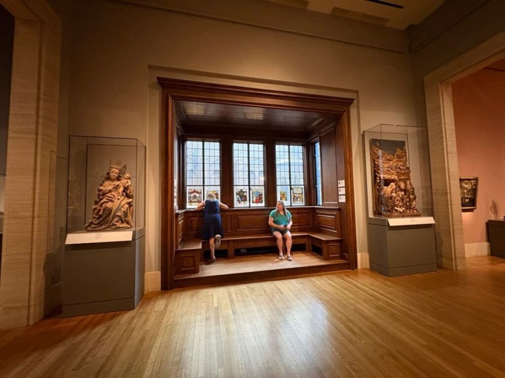 Two people sit on a wooden bench in an art museum alcove with artwork displayed on either side and natural light coming through the windows behind them.