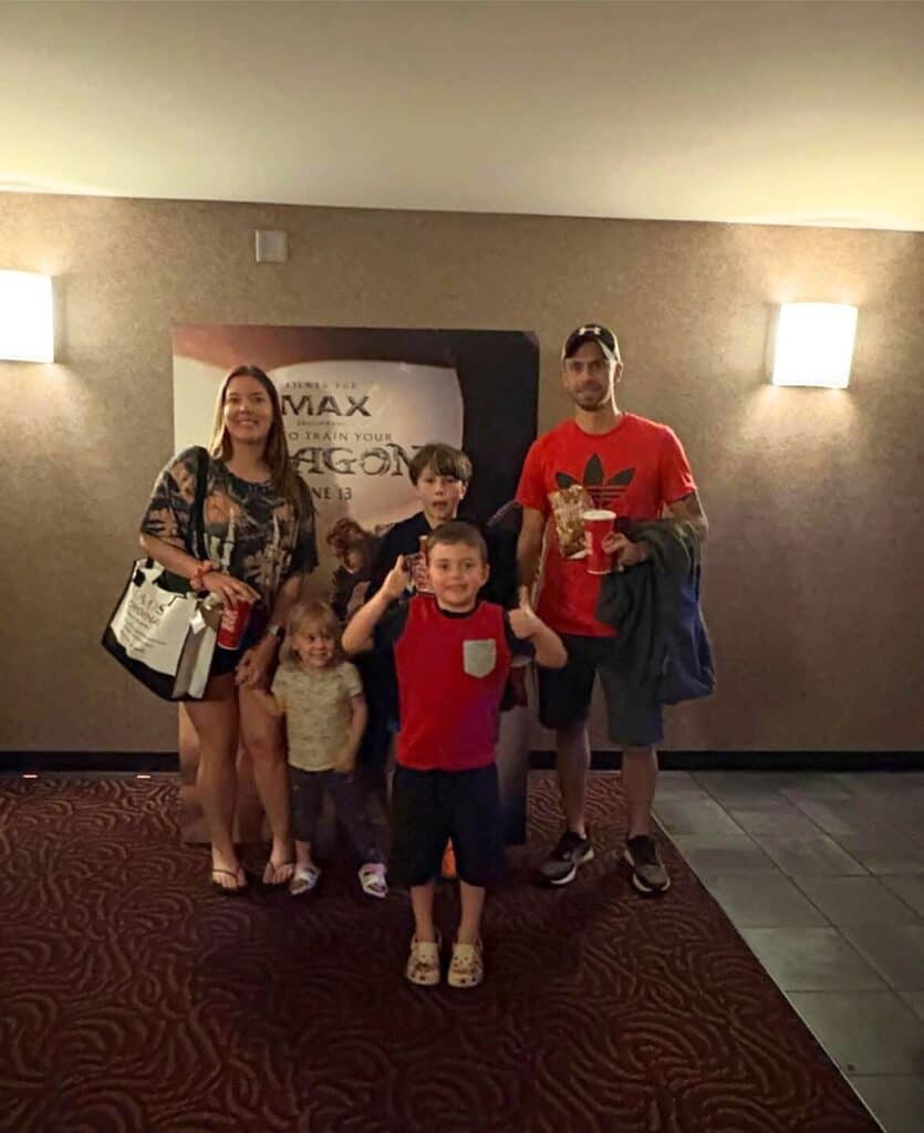 A family of five poses in a dimly lit hallway, with a movie poster in the background. The children stand in front, while the adults stand behind them.