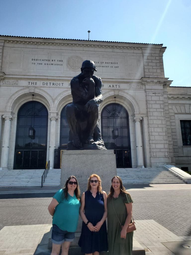 Three women stand in front of the “The Thinker” sculpture outside the Detroit Institute of Arts on a sunny day.