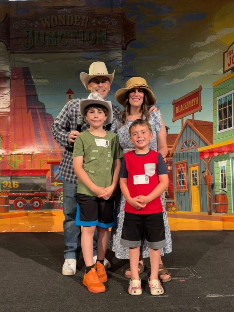 A man, woman, and two boys stand smiling in front of a Western-themed backdrop with buildings and signs, all wearing hats and casual summer clothing.
