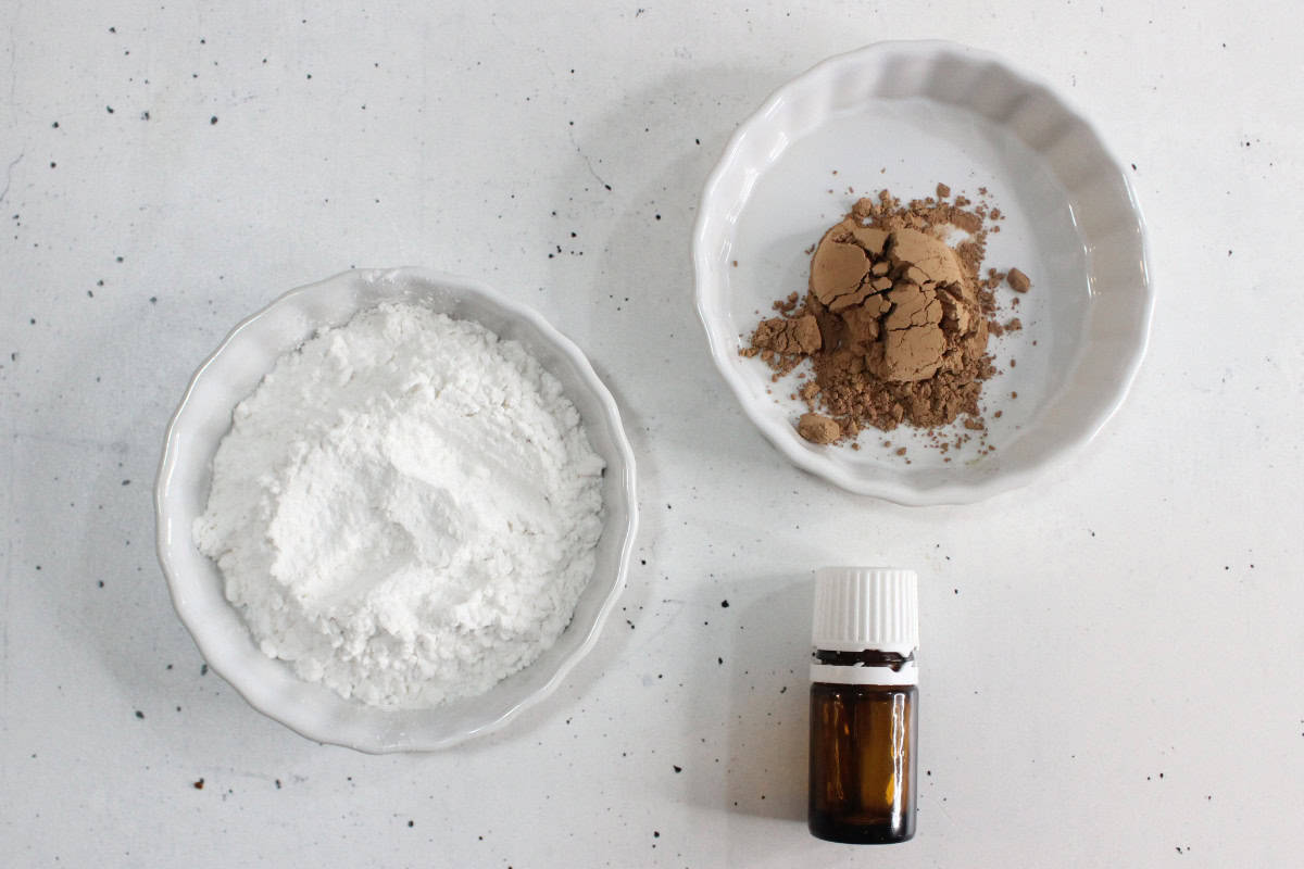 Two white dishes on a white surface, one with white powder, the other with brown powder, sit next to a small amber glass bottle with a white cap—perfect ingredients to make DIY dry shampoo at home.