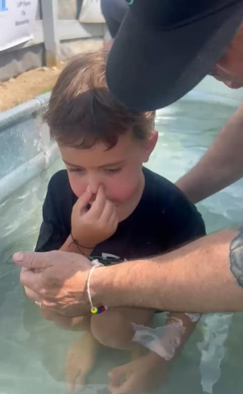 A child sits in a shallow pool, pinching their nose, while an adult supports them by the arm, preparing for immersion amid the cheerful chaos of the day.