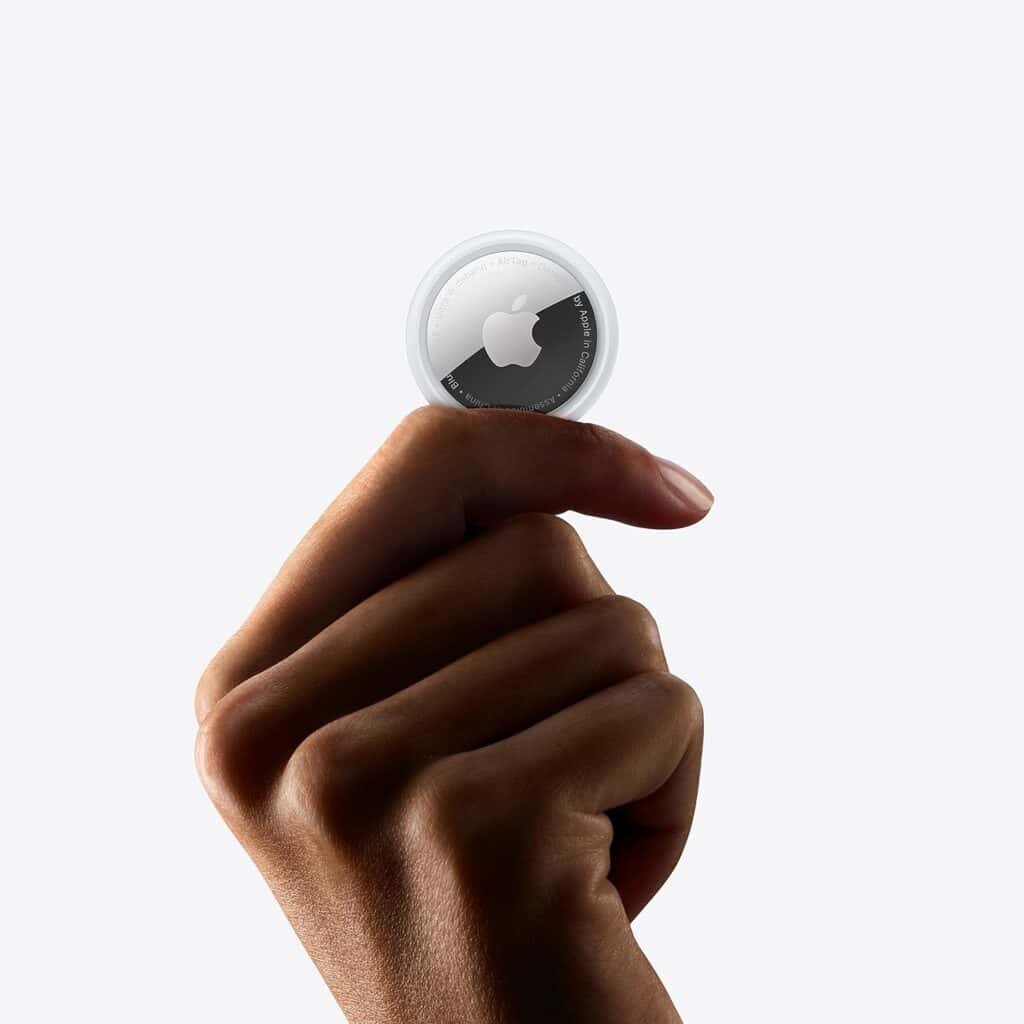A hand is holding an Apple AirTag between the thumb and index finger against a plain white background, showcasing one of the many ways to use Apple AirTags for tracking your valuables.