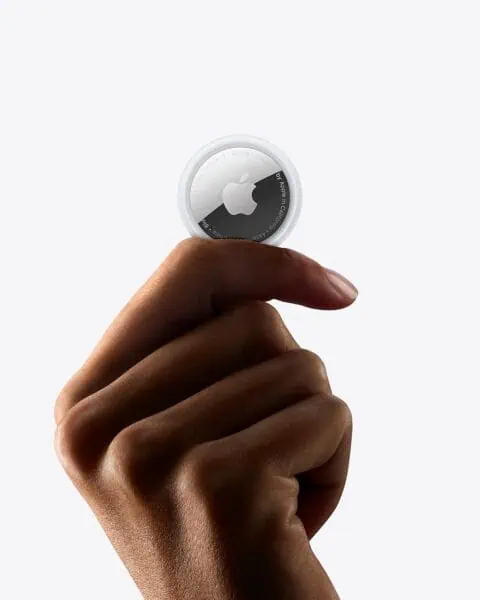 A hand is holding an Apple AirTag between the thumb and index finger against a plain white background, showcasing one of the many ways to use Apple AirTags for tracking your valuables.