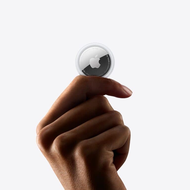 A hand is holding an Apple AirTag between the thumb and index finger against a plain white background, showcasing one of the many ways to use Apple AirTags for tracking your valuables.