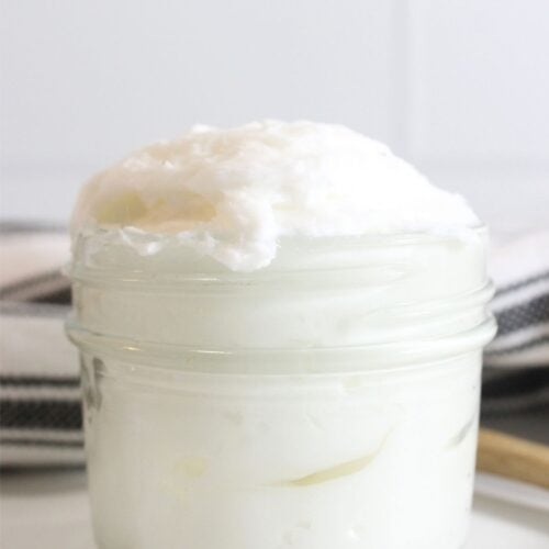 A small glass jar filled with a white, fluffy, whipped tallow sits on a white surface, with a blurred striped cloth in the background.