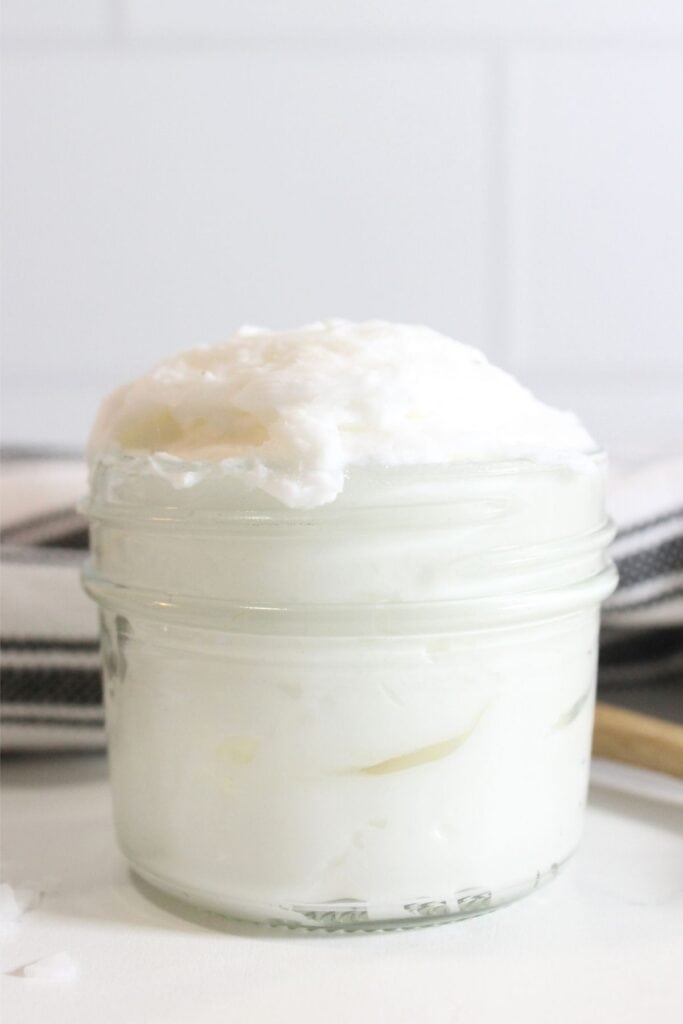 A small glass jar filled with a white, fluffy, whipped tallow sits on a white surface, with a blurred striped cloth in the background.