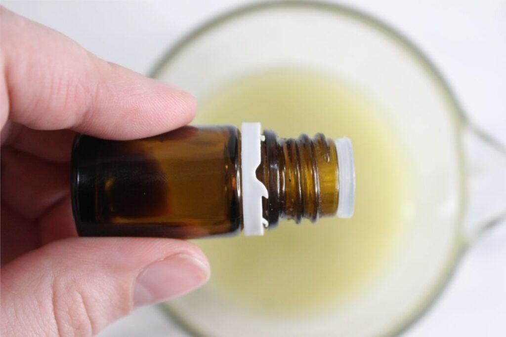 A hand holds a small amber glass bottle with a white cap above a glass bowl of light yellow liquid, ready to make homemade deodorant.