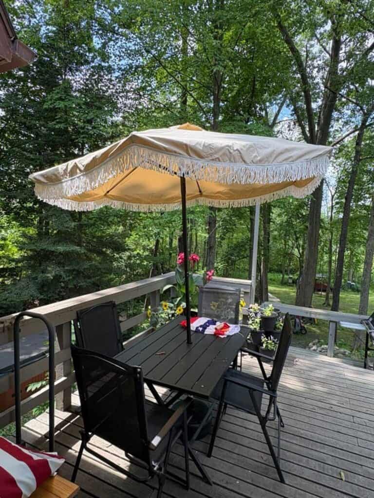 A patio table with four black chairs and a beige fringed umbrella on a wooden deck, surrounded by potted flowers and trees—ready for cake and Connor’s big news.