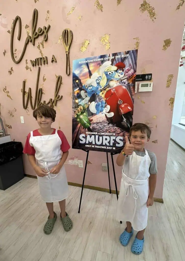 Two young boys wearing aprons stand in front of a "Smurfs" movie poster inside a bakery with a pink "Cakes with a Whisk" wall sign. Amid the cake chaos, one boy gives a thumbs up, ready for Connor’s Big News.
