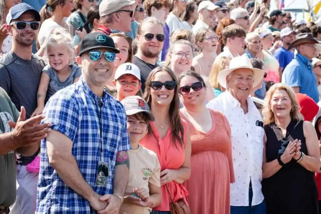A large group of people, including adults and children, stand outdoors in sunny weather, smiling and clapping while facing the camera—clearly celebrating Connor’s big news.