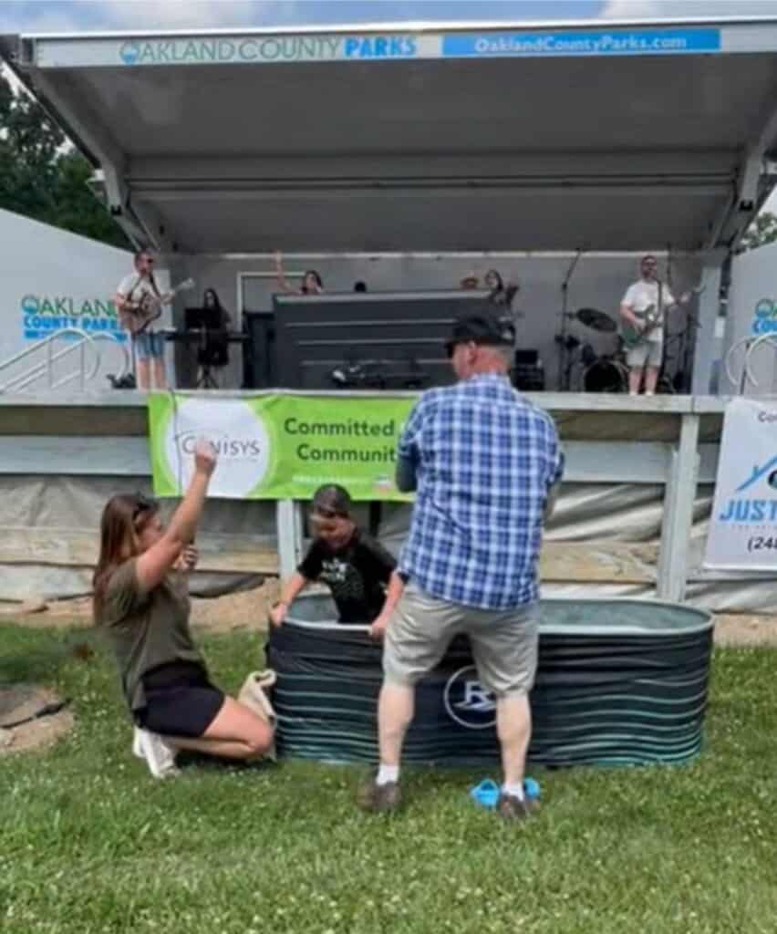 A woman kneels beside a stock tank while a man stands nearby; behind them, a band performs on stage at an outdoor community event buzzing with the excitement of Connor’s Big News.