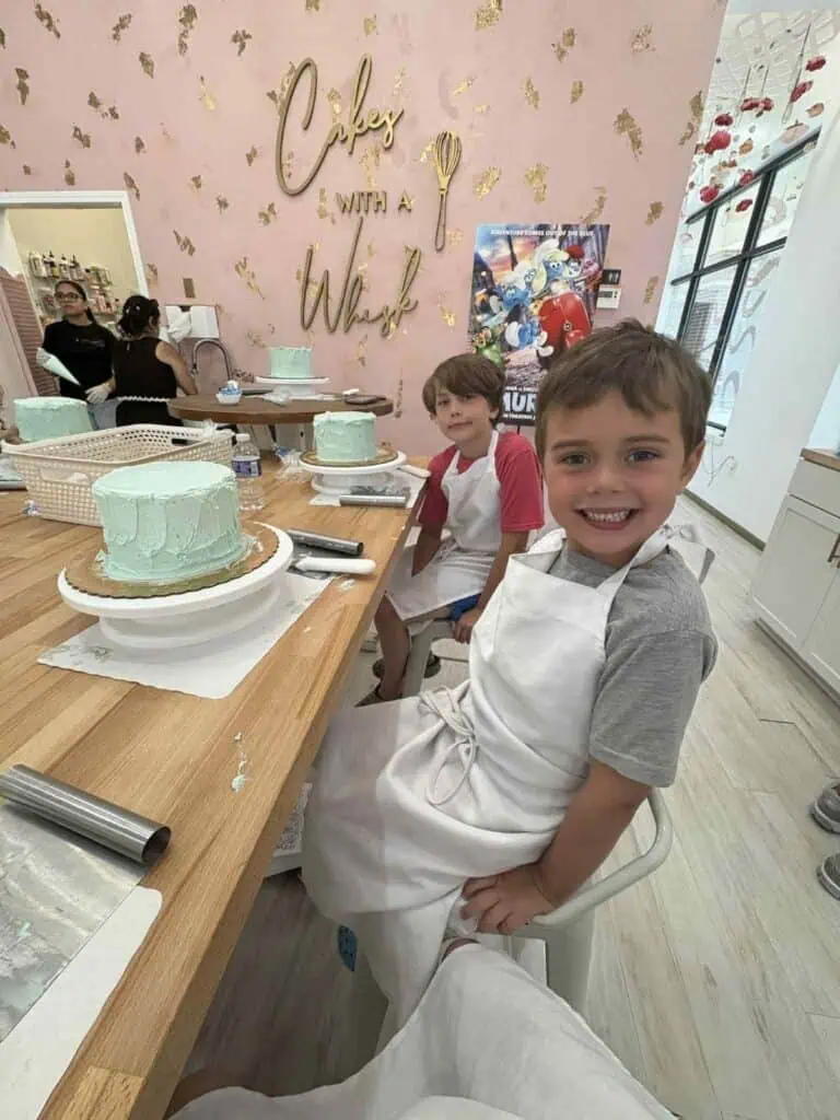 Connor and a friend, wearing aprons, sit at a table with partially decorated cakes in front of them, surrounded by pink walls and gold lettering—ready for some big news in this lively cake decorating studio.