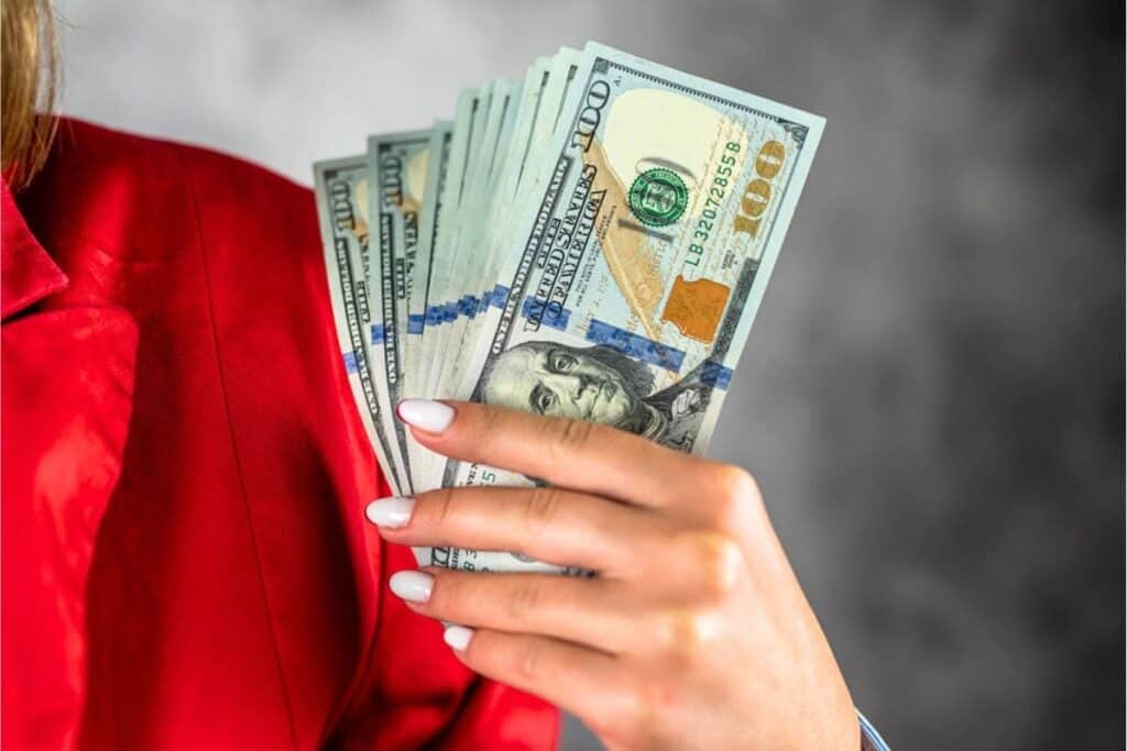A person wearing a red blazer holds several fanned-out U.S. one hundred dollar bills in their hand, symbolizing effective debt management and paying off loans with smart financial strategies.