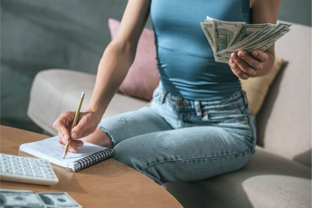 A person sits on a couch, counting cash in one hand while writing loan strategies in a notebook with the other; a calculator and more cash are on the table.