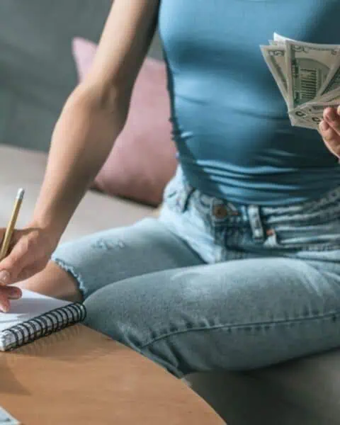 A person sits on a couch, counting cash in one hand while writing loan strategies in a notebook with the other; a calculator and more cash are on the table.