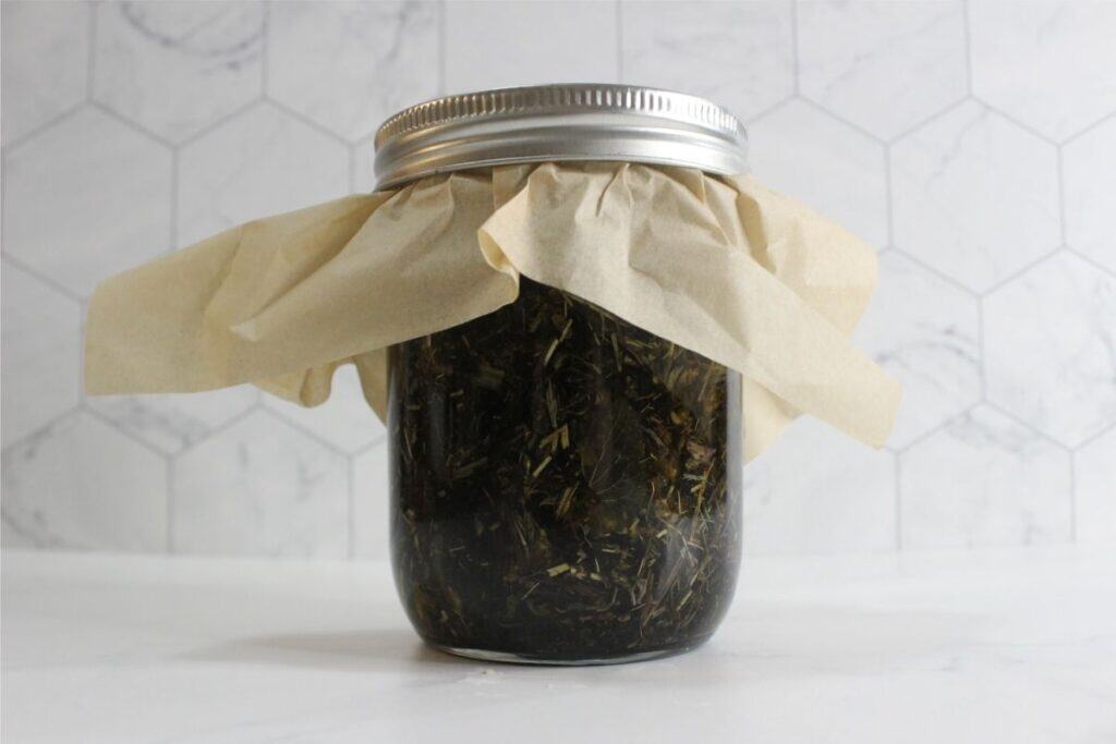 A glass jar filled with dried herbs—perfect for homemade deodorant—is covered with parchment paper and sealed with a metal lid, set against a white hexagonal tile background.