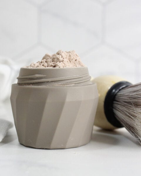 A beige container filled with a light brown DIY Dry Shampoo powder sits on a white surface next to a shaving brush with a cream-colored handle.