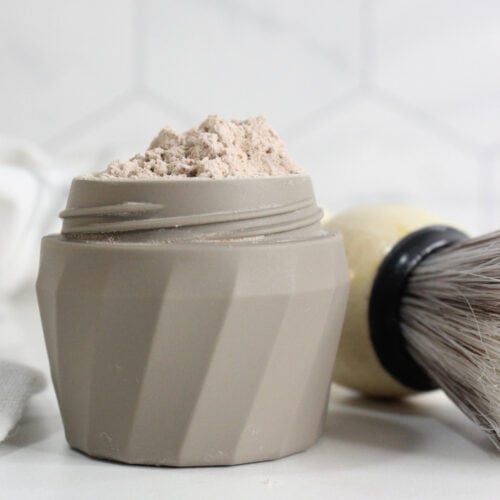 A beige container filled with a light brown DIY Dry Shampoo powder sits on a white surface next to a shaving brush with a cream-colored handle.