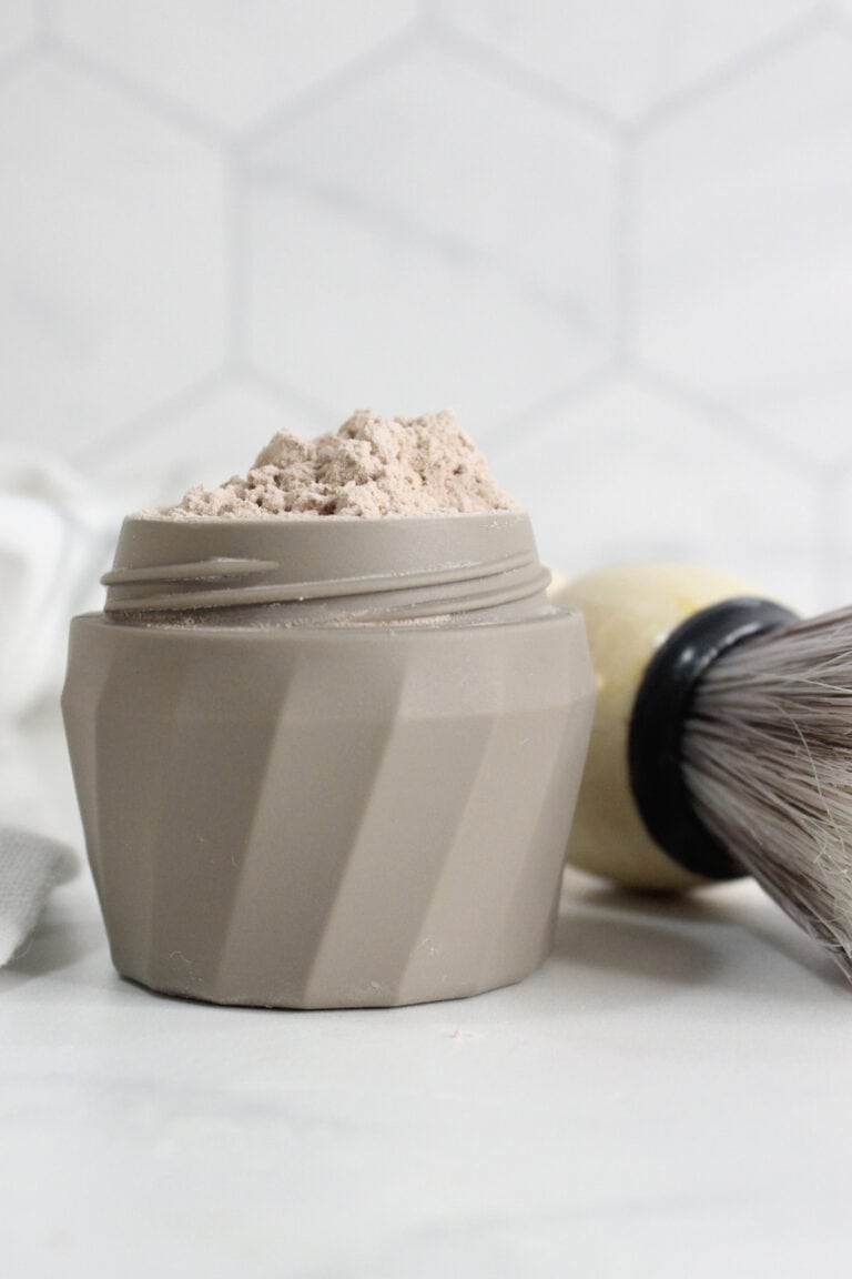 A beige container filled with a light brown DIY Dry Shampoo powder sits on a white surface next to a shaving brush with a cream-colored handle.