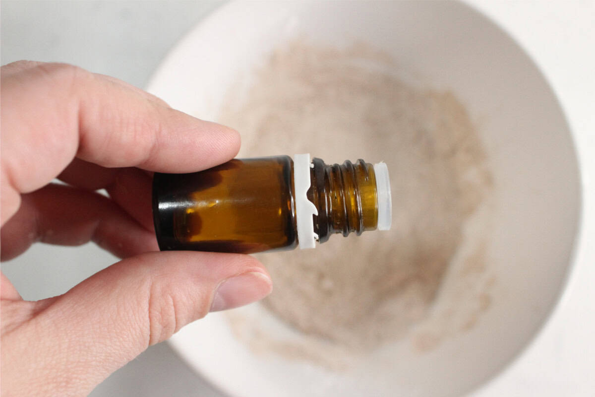 A hand holds a small brown glass bottle above a white bowl containing a powdery mixture, ready to make DIY dry shampoo.