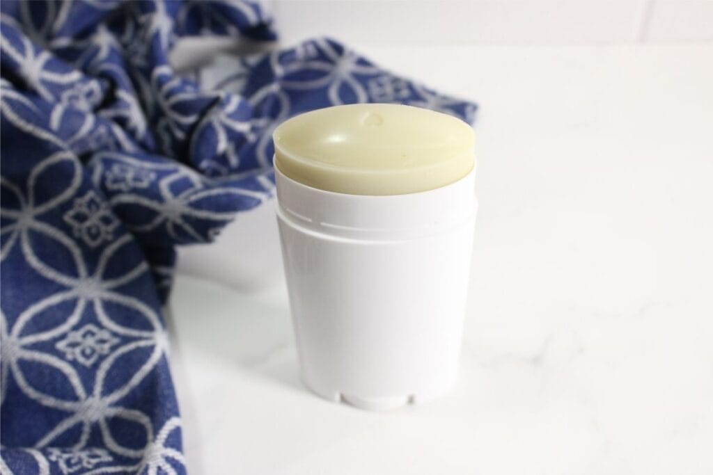A white homemade deodorant stick with a pale yellow top sits on a white surface next to a blue patterned cloth.