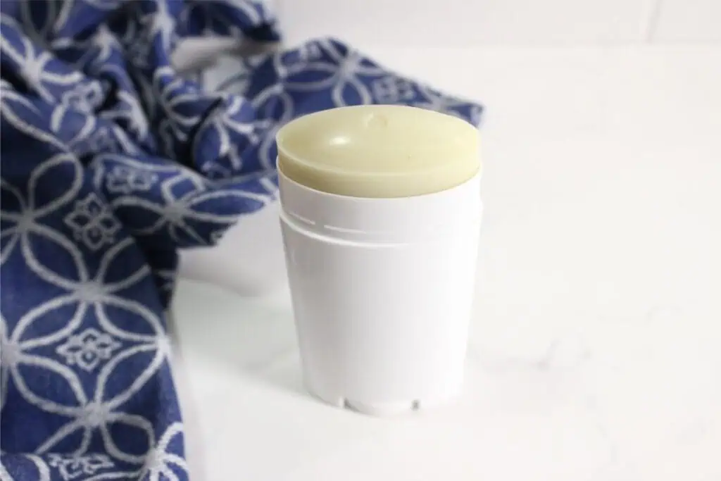 A white homemade deodorant stick with a pale yellow top sits on a white surface next to a blue patterned cloth.