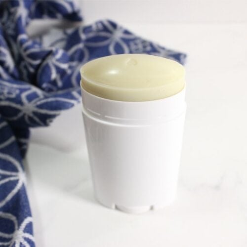 A white homemade deodorant stick with a pale yellow top sits on a white surface next to a blue patterned cloth.