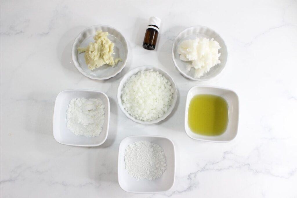 Six small dishes and one small bottle are arranged on a white surface, containing various ingredients like oils, powders, and chopped solids—perfect for those looking to make deodorant or try a homemade deodorant recipe.