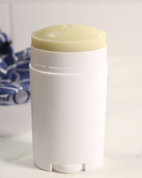 A white cylindrical homemade deodorant stick with an exposed yellowish top sits on a white surface, with part of a blue and white cloth in the background.