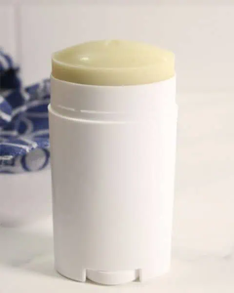 A white cylindrical homemade deodorant stick with an exposed yellowish top sits on a white surface, with part of a blue and white cloth in the background.