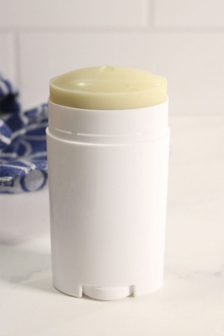 A white cylindrical homemade deodorant stick with an exposed yellowish top sits on a white surface, with part of a blue and white cloth in the background.