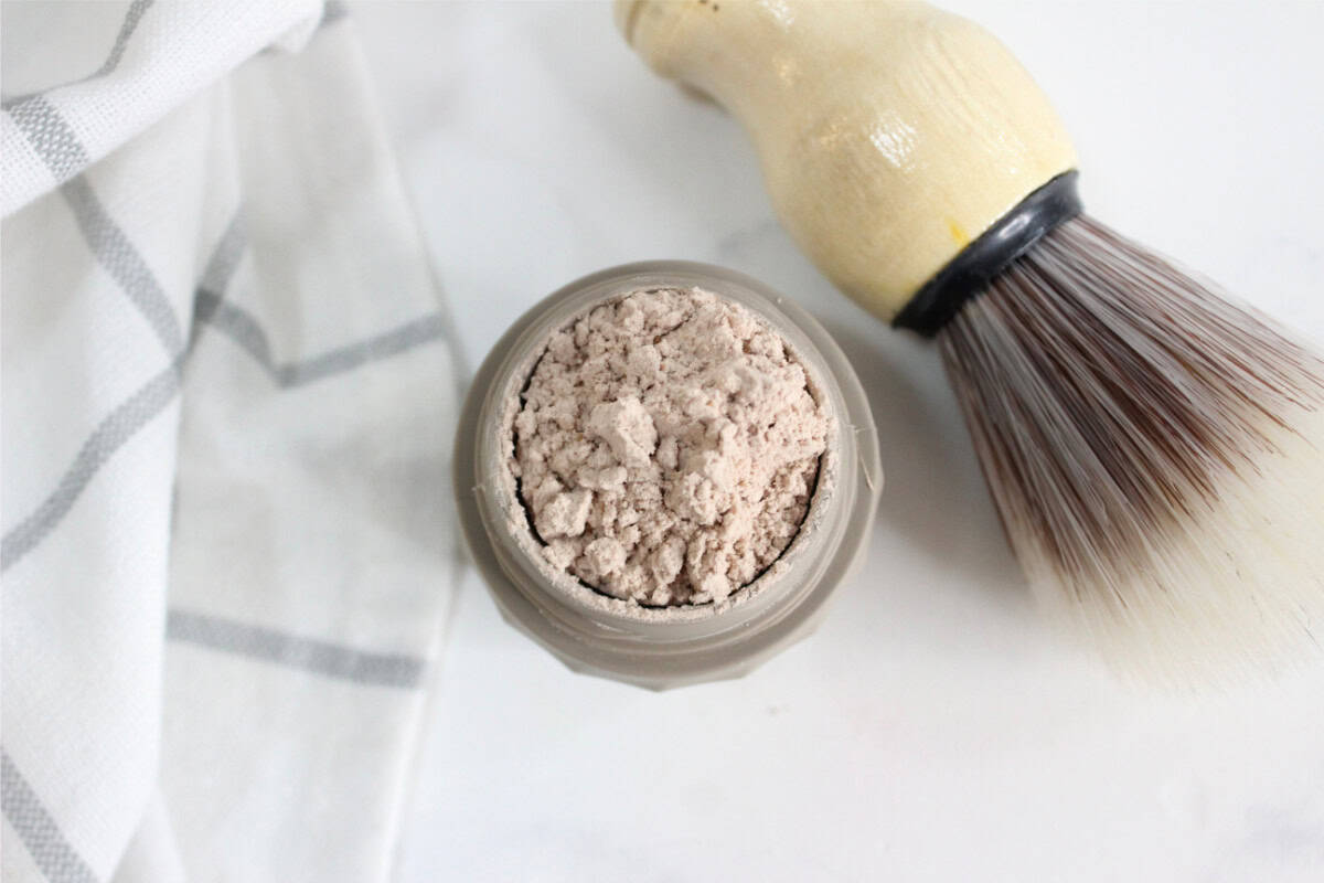 A jar of powdered shaving soap sits next to a shaving brush and a white cloth with gray stripes on a white surface—perfect inspiration if you want to make dry shampoo or explore other DIY grooming essentials.