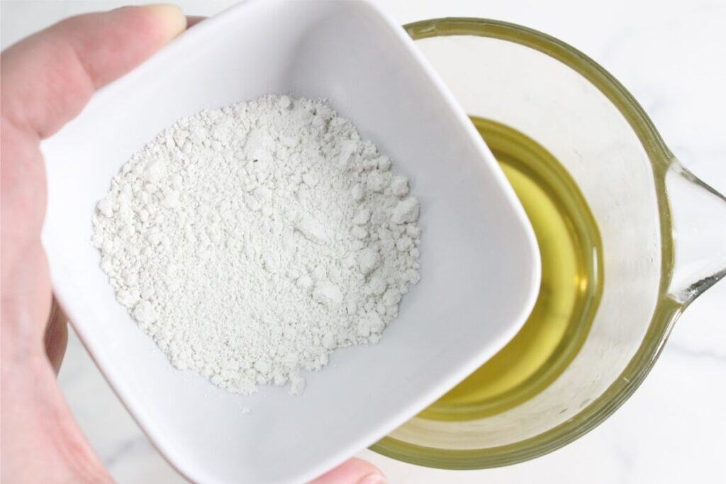 A hand holds a white bowl of white powder above a glass measuring cup filled with yellow liquid on a white surface, ready to make DIY deodorant.
