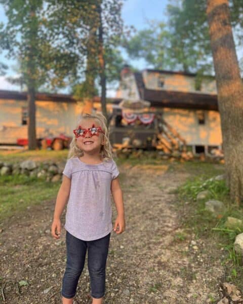 Young child wearing star-shaped red glasses stands on a gravel path in front of a house decorated with patriotic banners, surrounded by trees—unaware of Connor’s big news stirring up chaos inside.