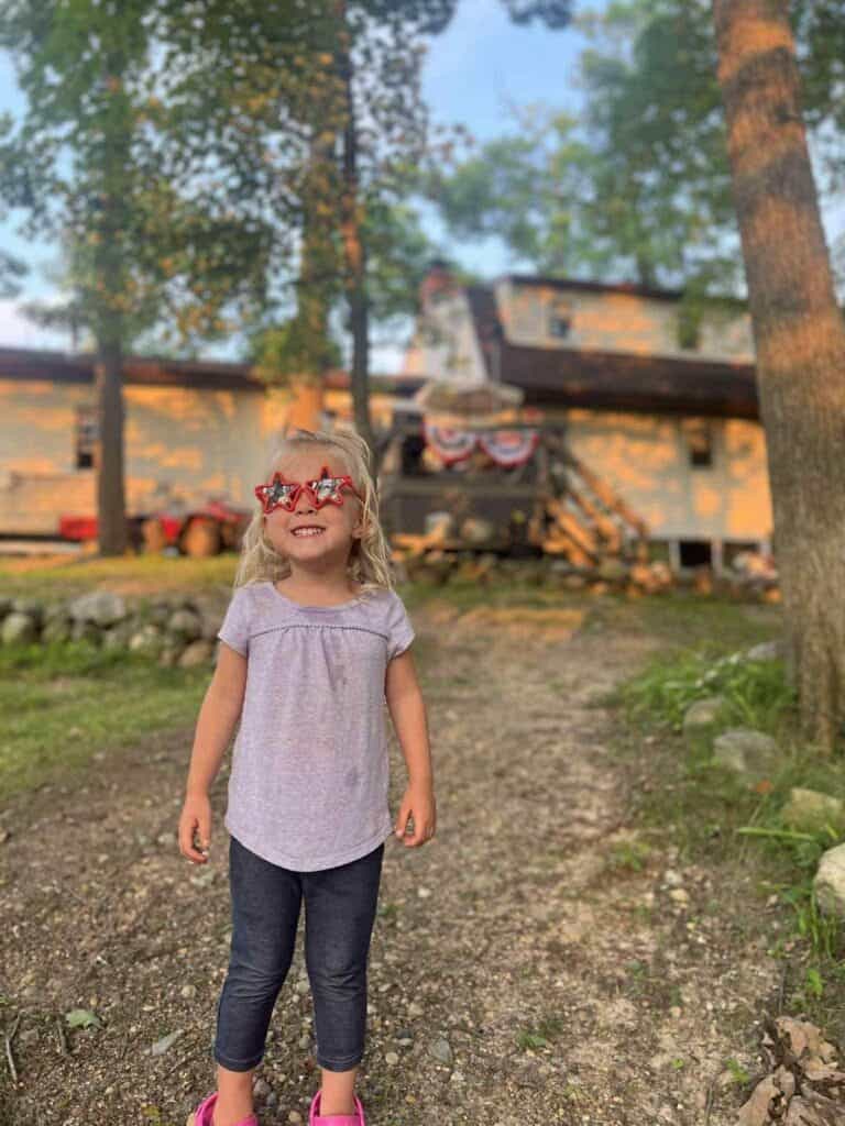 Young child wearing star-shaped red glasses stands on a gravel path in front of a house decorated with patriotic banners, surrounded by trees—unaware of Connor’s big news stirring up chaos inside.